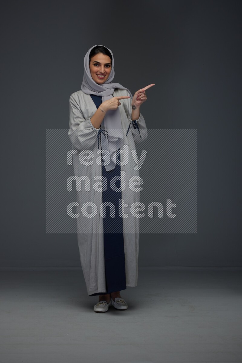 A Saudi woman wearing a light gray Abaya and head scarf standing and pointing in different directions eye level on a grey background
