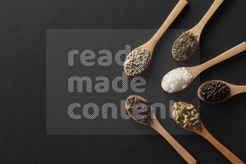 wooden spoons filled with white peppers, cloves, cardamom, salt, black peppers and basil on black flooring