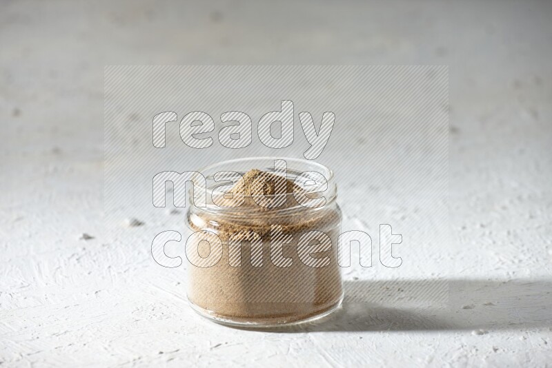A glass jar full of cumin powder on textured white flooring