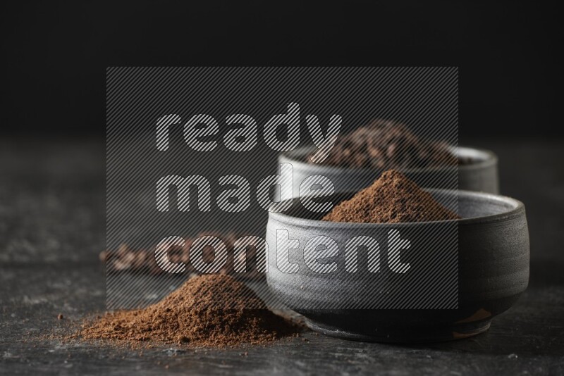 2 Black pottery bowls full of cloves and the other full of cloves powder on textured black flooring