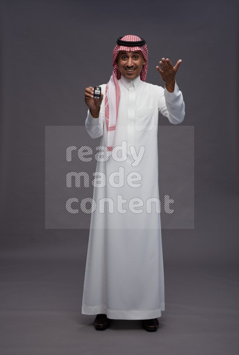Saudi man wearing thob and shomag standing holding key car on gray background