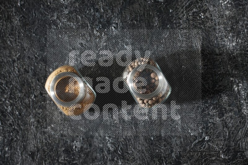 2 glass spice jars full of allspice powder and whole balls on a textured black flooring