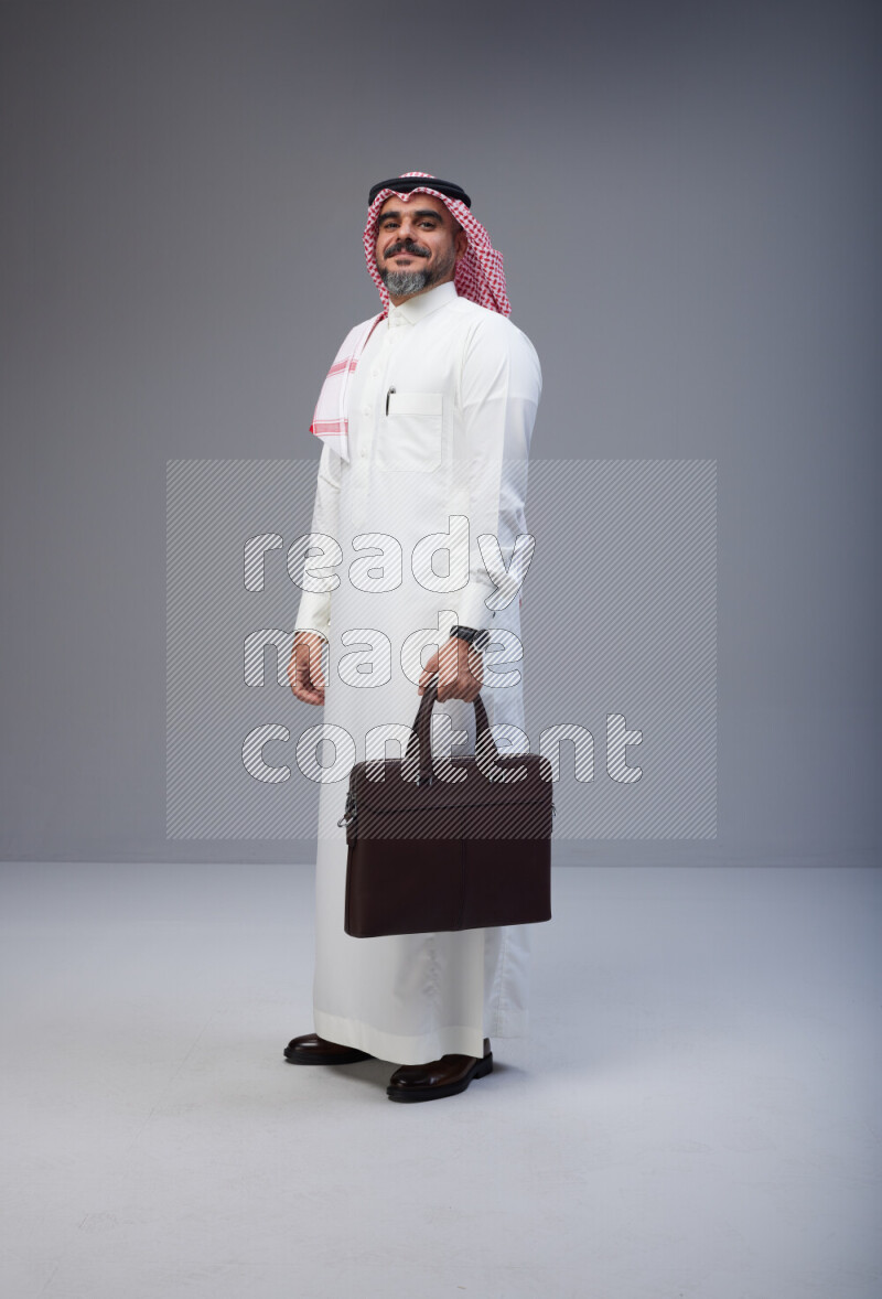 Saudi man Wearing Thob and red Shomag standing holding bag on Gray background