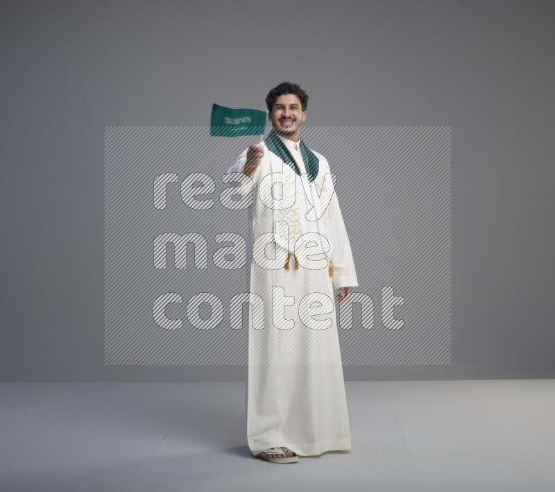 A Saudi man standing wearing thob and saudi flag scarf and holding small saudi flag on gray background