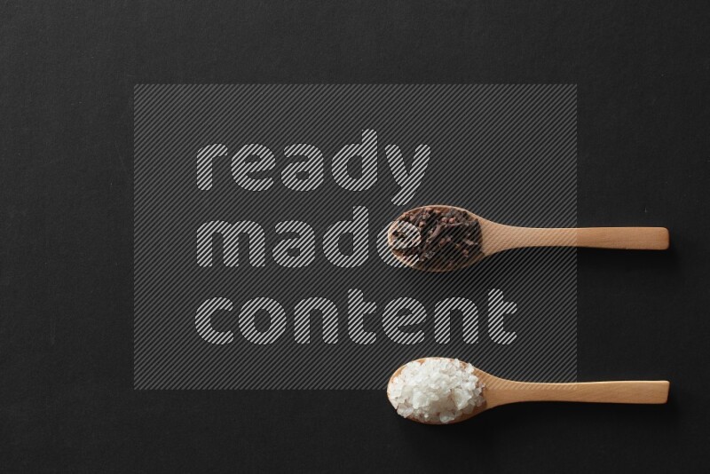 wooden spoons filled with white peppers, cloves, cardamom, salt, black peppers and basil on black flooring