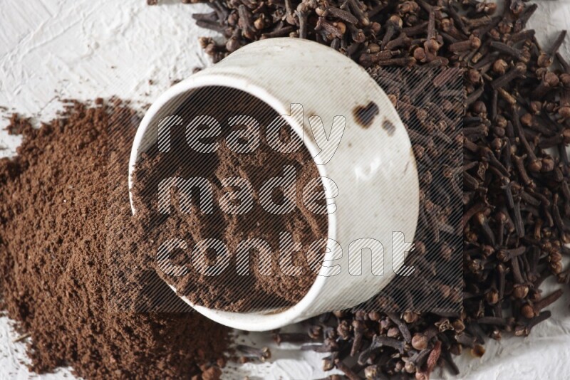 وعاء بيج من السيراميك ممتلئ ببودرة القرنفل وبعض بودرة القرنفل سقطت بخارجه على أرضية بيضاء