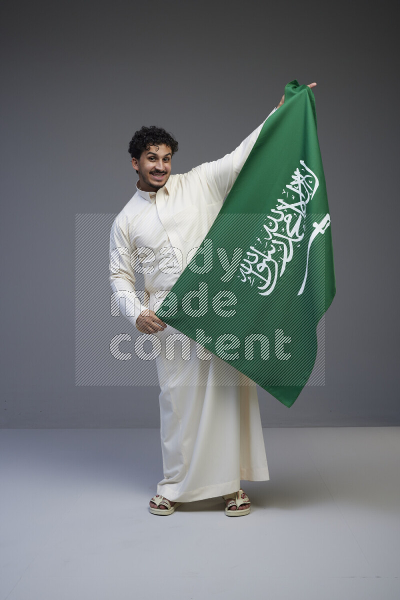 A Saudi man standing wearing thob holding big Saudi flag on gray background