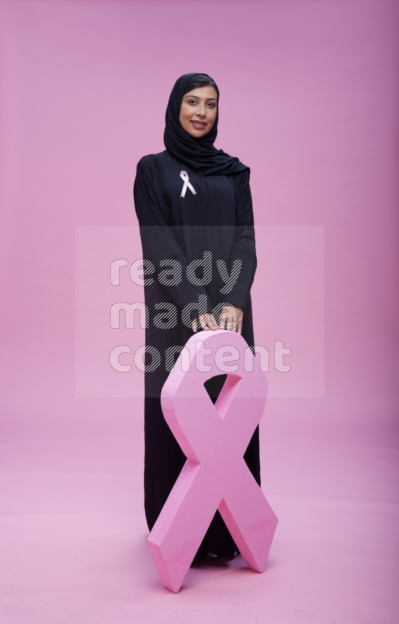 Saudi woman wearing pink ribbon on abaya standing holding awareness ribbon on plain pink background