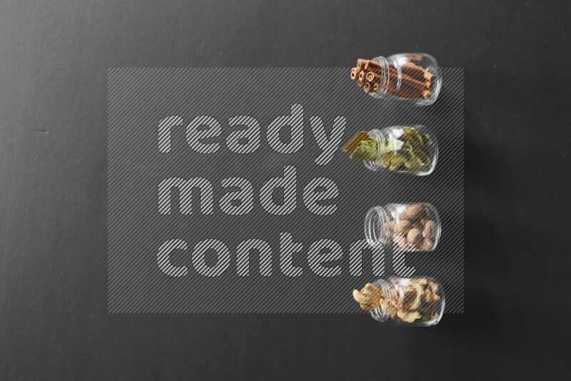 4 glass spice jars full of laurel bay leaves, ginger, cinnamon and nutmeg on black flooring