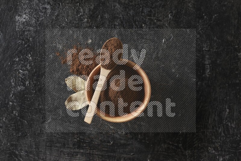 A wooden bowl and a wooden spoon full of cloves powder with laurel leaves on a textured black flooring