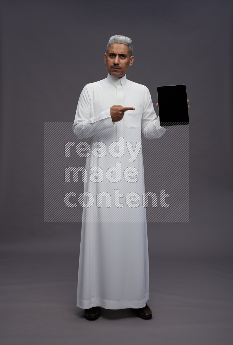 Saudi man wearing thob standing showing tablet to camera on gray background