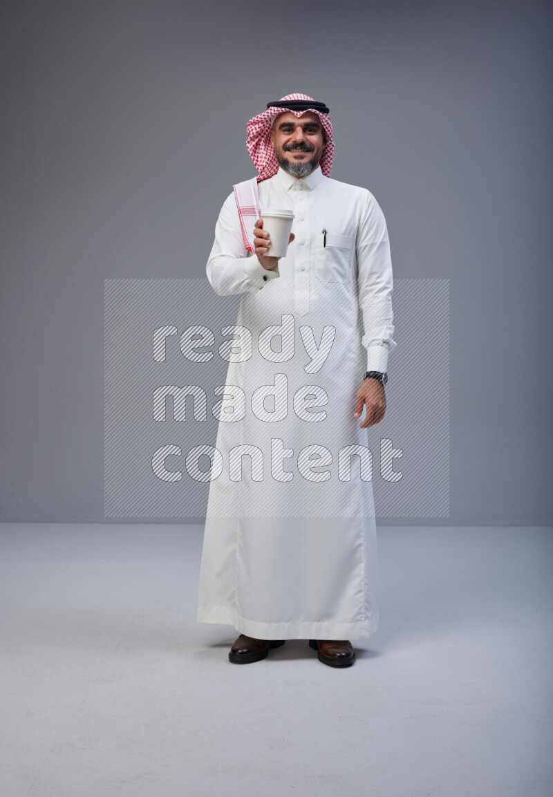 Saudi man Wearing Thob and red Shomag standing holding paper cup on Gray background