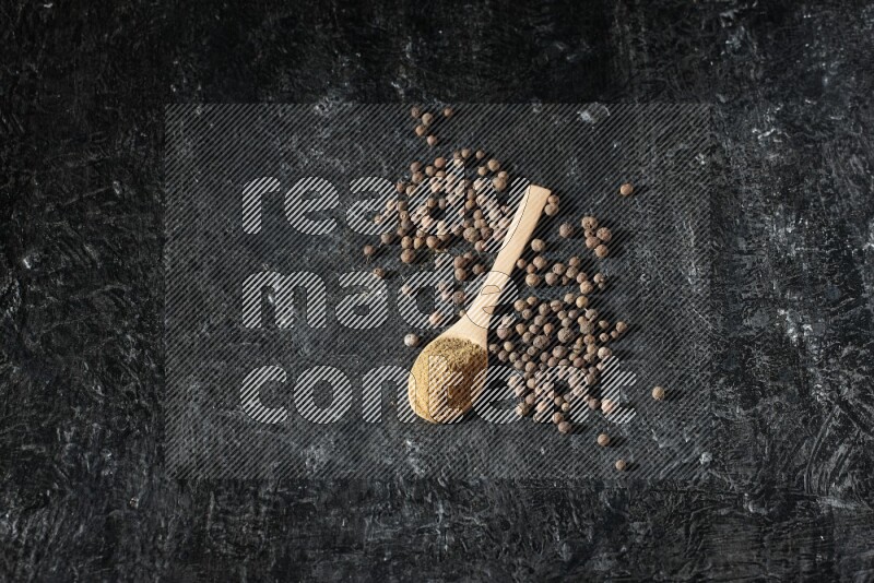 Wooden spoons full of all spice powder and allspice whole balls beside it on a textured black flooring