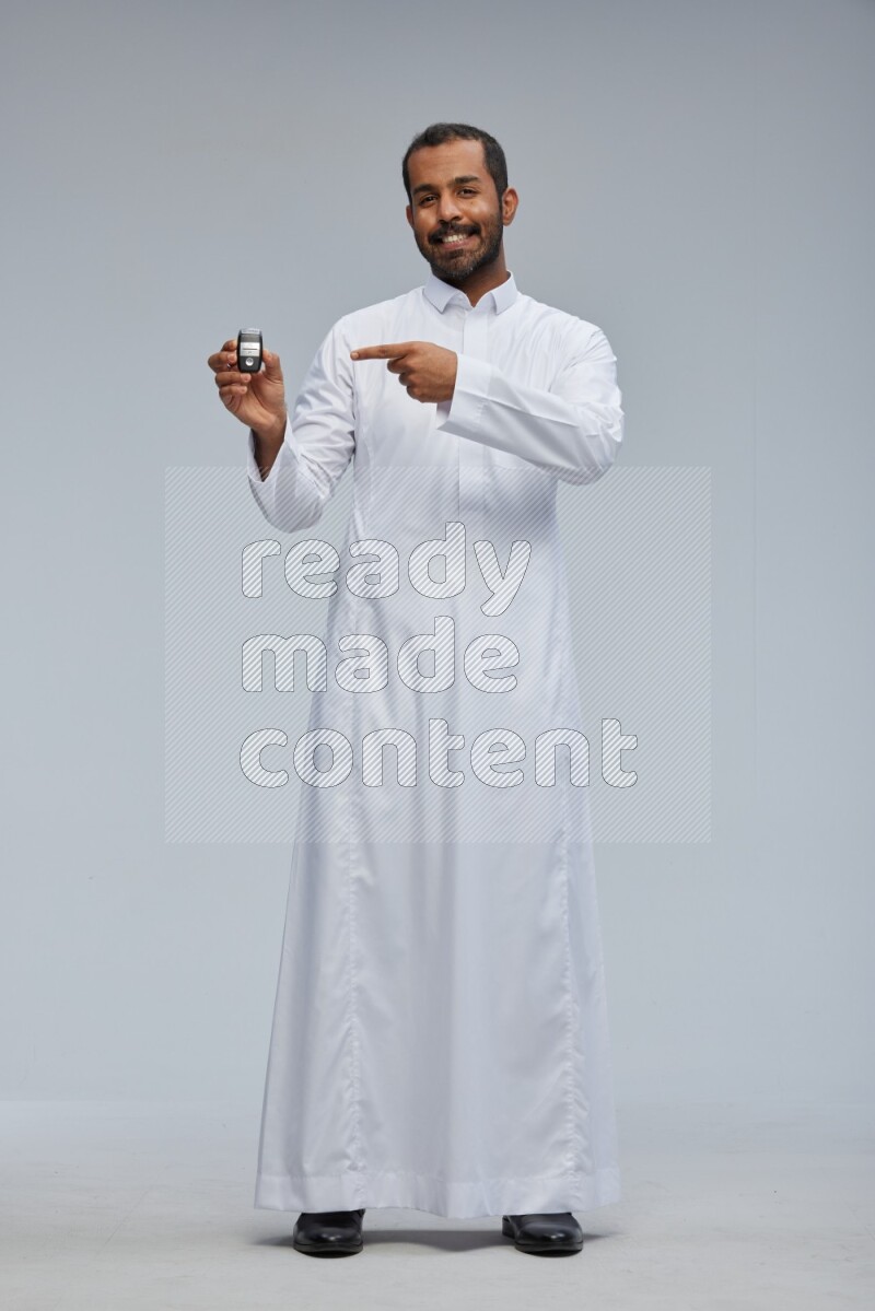 Saudi man wearing thob and shomag standing holding car key on gray background