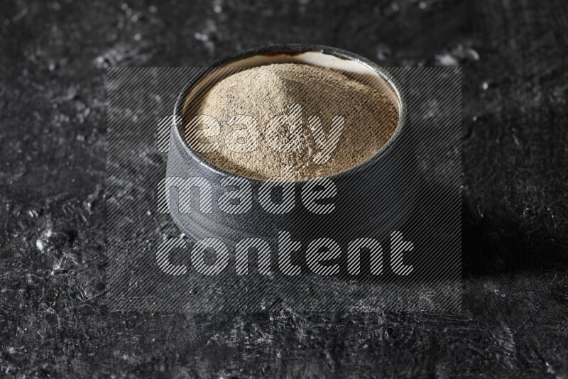 Black pottery bowl full of white pepper powder on textured black flooring