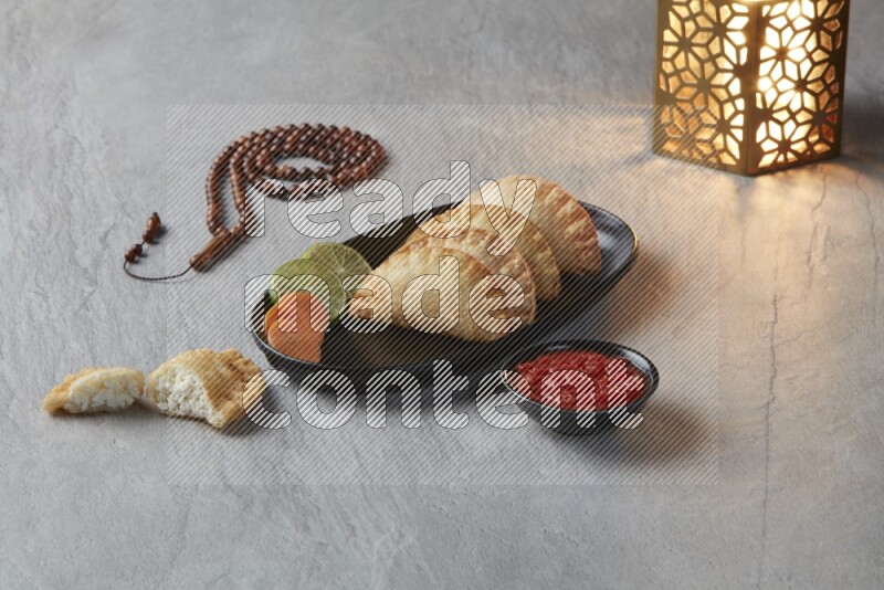 Four fried sambosas in an oval shaped black plate, beside a cut cheese sambosa, a brown misbaha and a golden lantern on a gray background