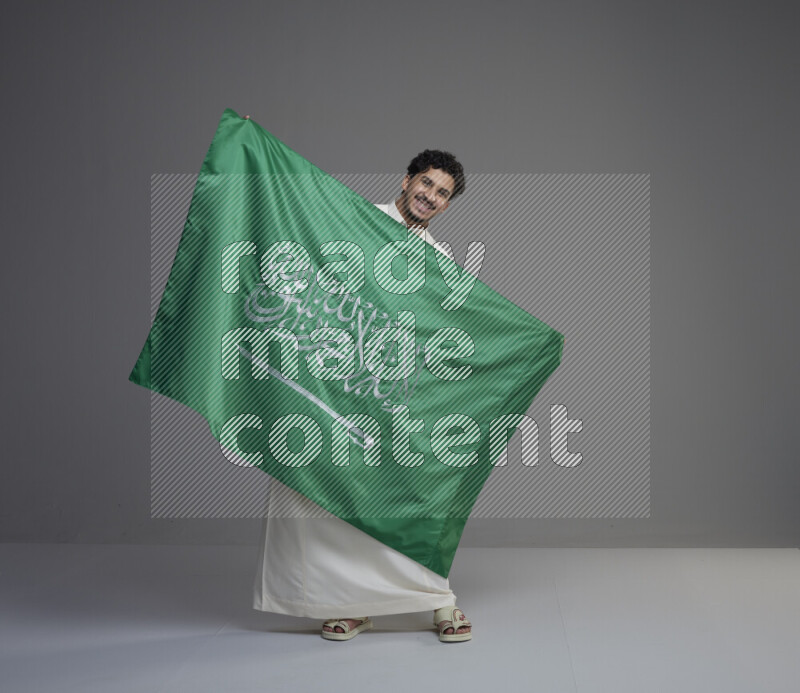 A saudi man standing wearing thob holding big saudi flag on gray background