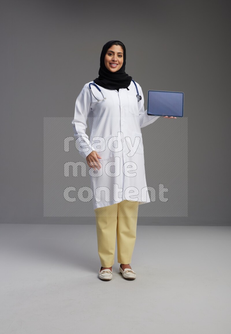 Saudi woman wearing lab coat with stethoscope standing showing tablet to camera with sign in the back on Gray background