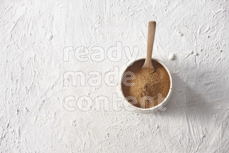 Ceramic beige bowl full of cinnamon powder with a wooden spoon on a textured white background