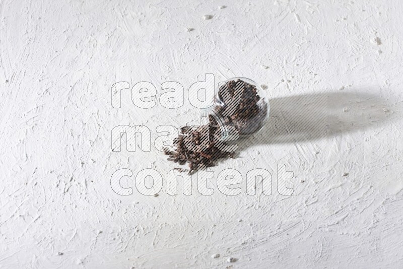 A flipped glass spice jar full of cloves and cloves came out of it on textured white flooring