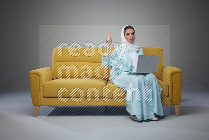 A Saudi woman sitting on a yellow Sofa With her Laptop Pointing wearing Blue Abaya