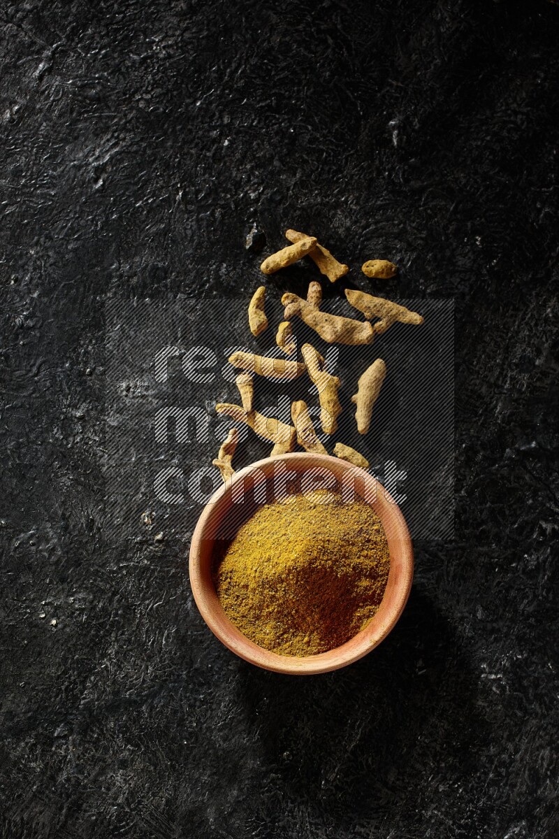 A wooden bowl full of turmeric powder with dried turmeric fingers on textured black flooring