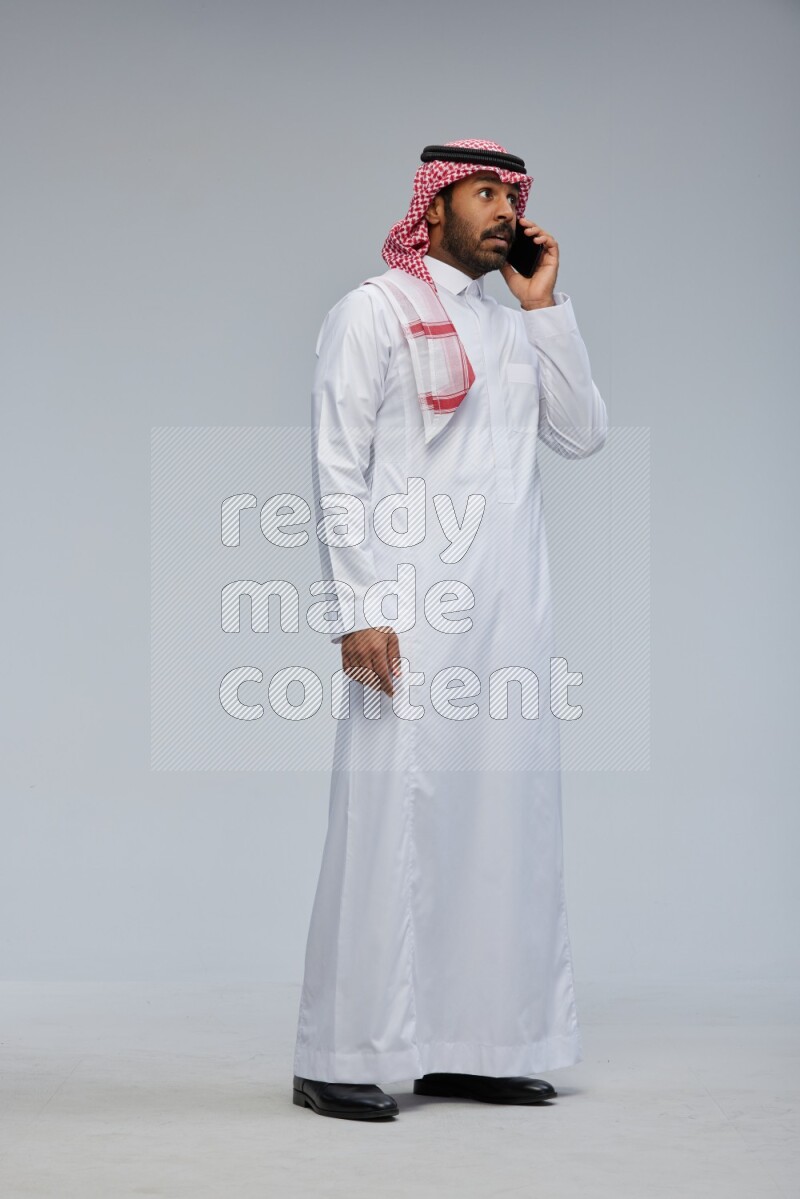 Saudi man Wearing Thob and shomag standing talking on phone on Gray background