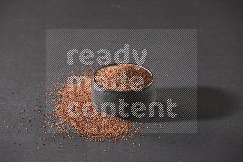 A black pottery bowl full of garden cress seeds surrounded by seeds on a black flooring