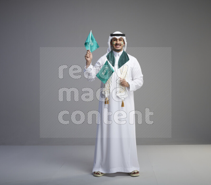 A saudi man standing wearing thob and white shomag with face painting and saudi flag scarf and holding small saudi flag on gray background