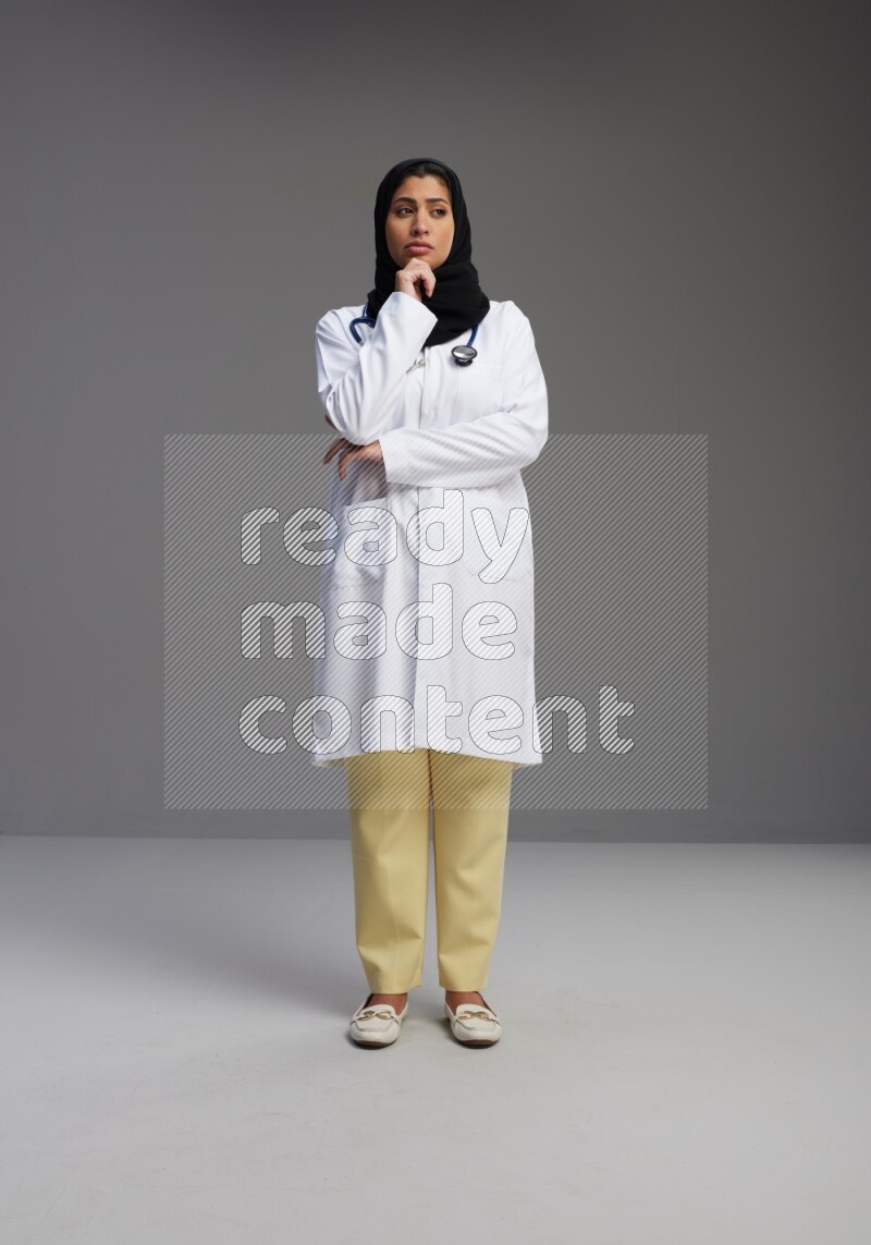 Saudi woman wearing lab coat with stethoscope standing interacting with the camera on Gray background