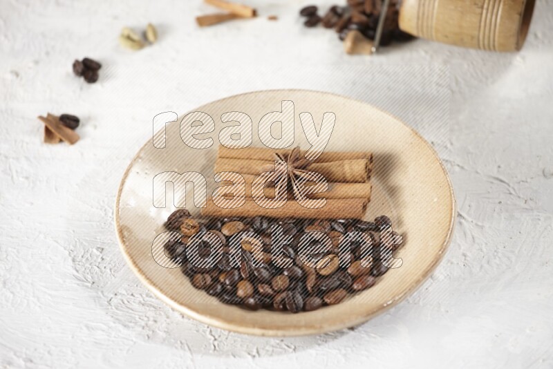Beige plate full of coffee beans, cinnamon sticks and star anise with a coffee grinder, coffee beans, cinnamon pieces and cardamom next of it on white background