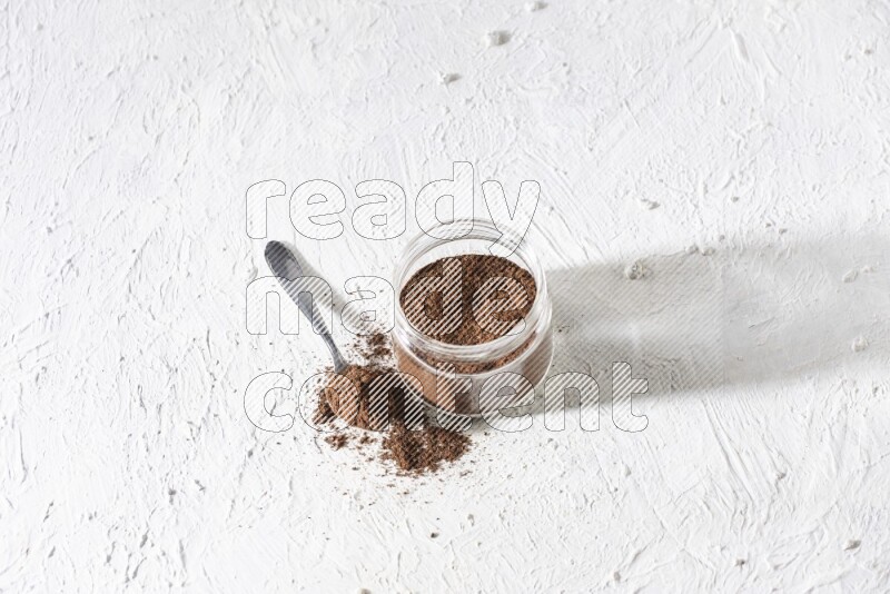 A glass jar full of cloves powder with a metal spoon on a textured white flooring