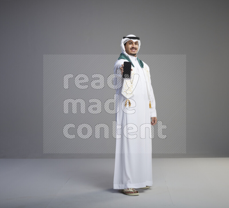 A Saudi man standing wearing thob and white shomag with flag scarf on his neak showing phone on gray background