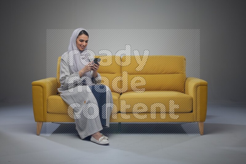 A Saudi woman wearing a light gray Abaya and white head scarf sitting on a yellow sofa and using her phone eye level on a grey background