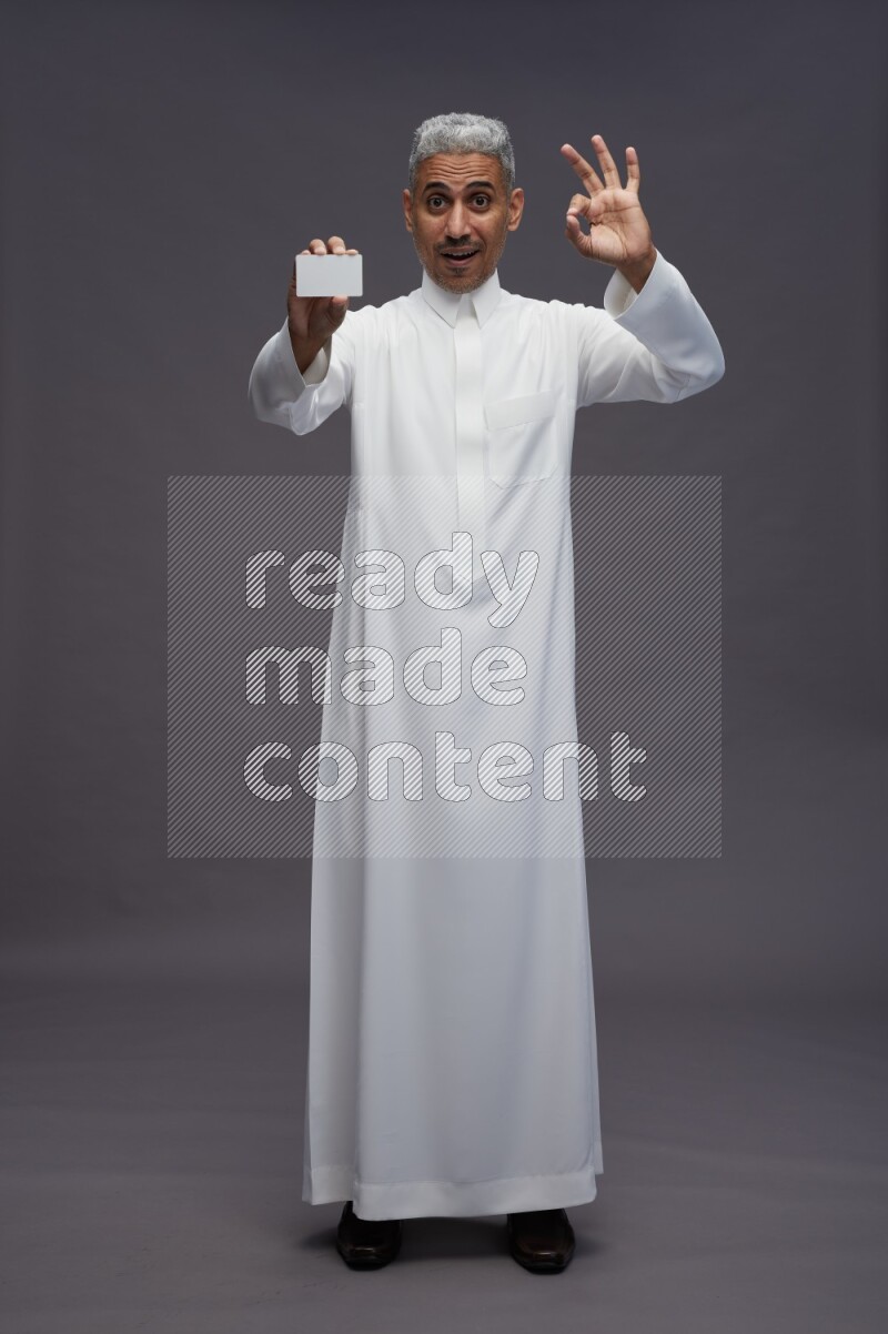 Saudi man wearing thob standing holding ATM card on gray background