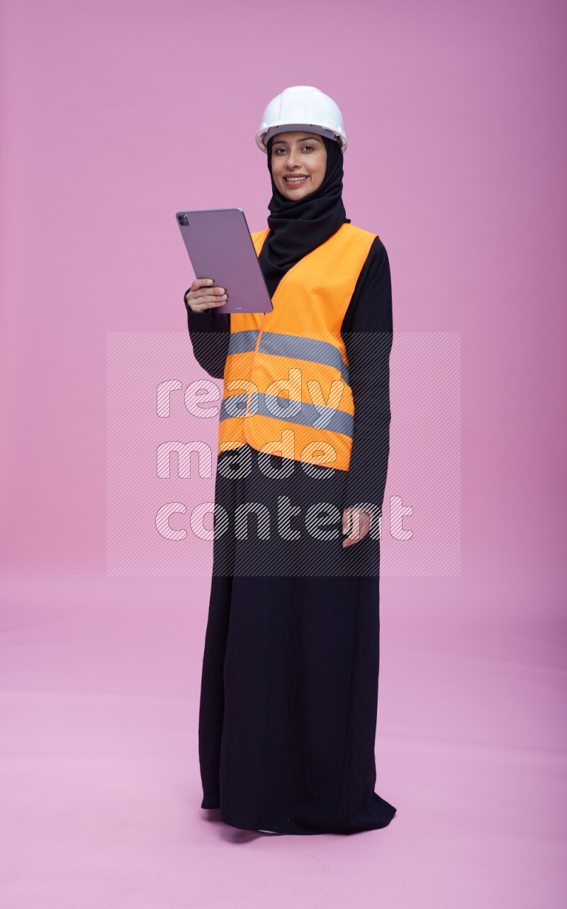 Saudi woman wearing Abaya with engineer vest and helmet standing working on tablet on pink background