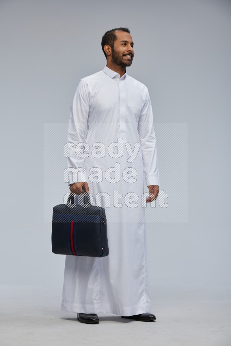 Saudi man wearing thob standing holding suitcase on gray background