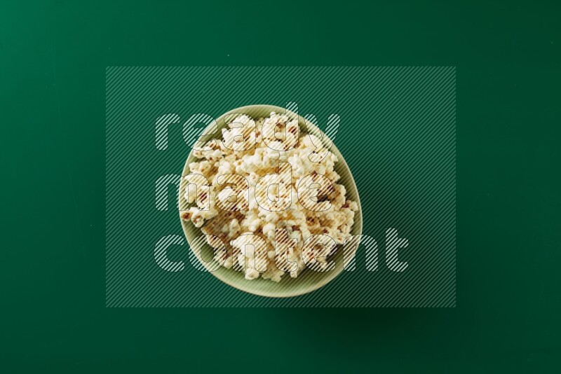 A green ceramic bowl full of popcorn on a green background in a top view shot