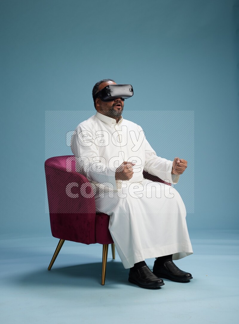 Saudi Man without shimag sitting on chair  gaming with VR on blue background
