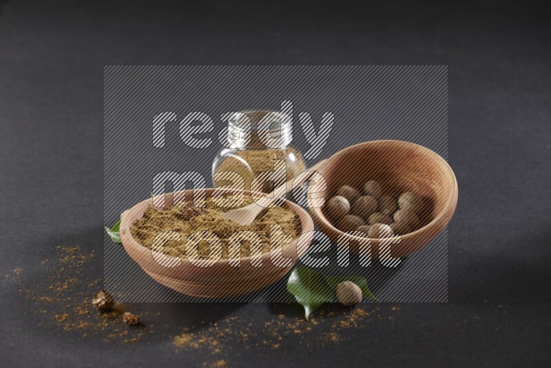 A wooden bowl with a spoon in it and glass spice jar all, full of nutmeg powder and a wooden bowl full of whole nutmeg seeds on a black flooring