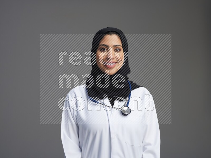 Saudi woman wearing lab coat with stethoscope standing interacting with the camera on Gray background