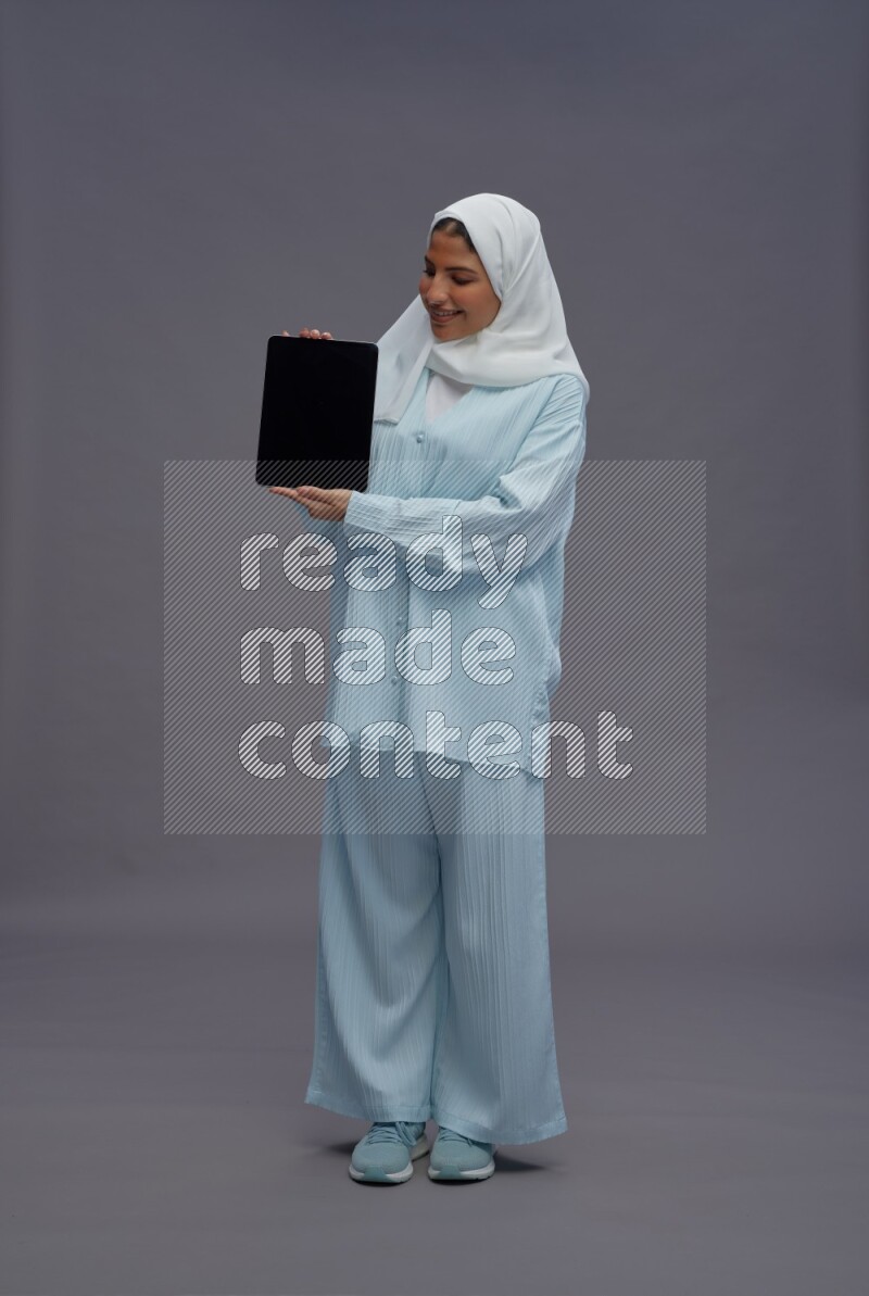 Saudi woman wearing hijab clothes standing showing tablet to camera on gray background