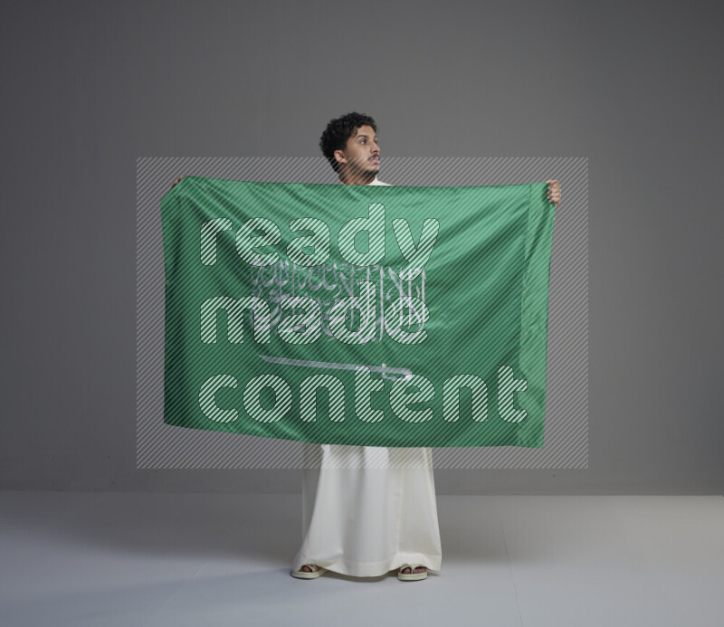 A Saudi man standing wearing thob holding big Saudi flag on gray background
