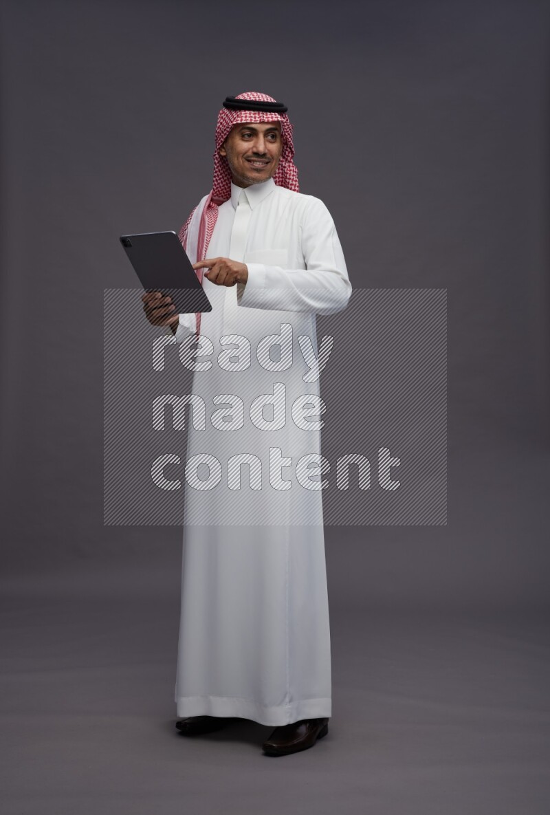 Saudi man wearing thob and shomag standing working on tablet on gray background