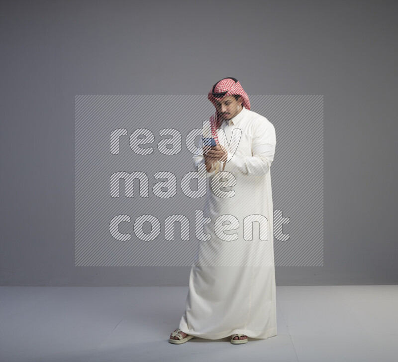 A  Saudi man standing wearing thob and red shomag texting on phone on gray background