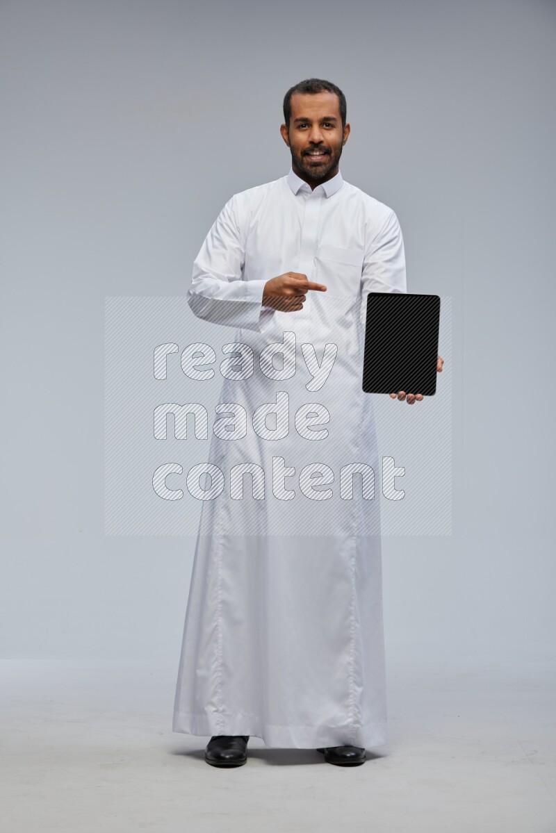 Saudi man wearing Thob standing showing tablet to camera on Gray background