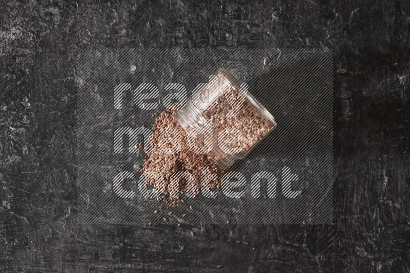A glass jar full of flaxseeds flipped and seeds spread out on a textured black flooring