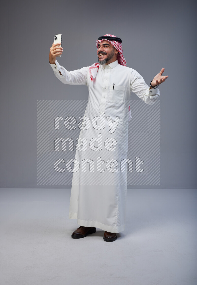 Saudi man Wearing Thob and red Shomag standing taking selfie on Gray background