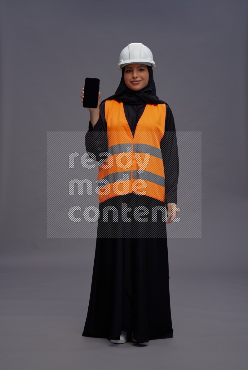 Saudi woman wearing Abaya with engineer vest standing showing phone to camera on gray background