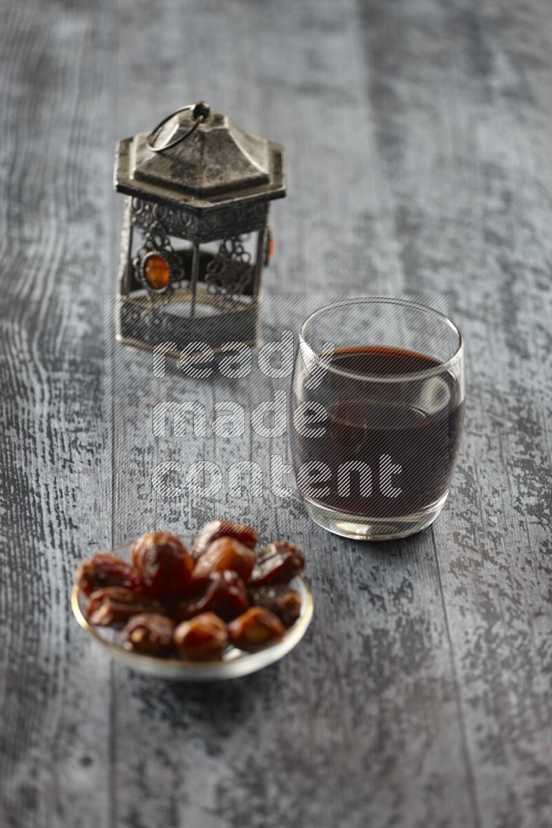 A silver lantern with different drinks, dates, nuts, prayer beads and quran on grey wooden background